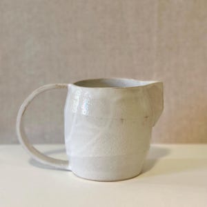 May include: A handmade, off-white ceramic pitcher with a rounded handle and a spout. The pitcher has a textured surface and a slightly irregular shape, suggesting a rustic or artisanal aesthetic. The neutral color and simple design make it suitable for various kitchen styles.