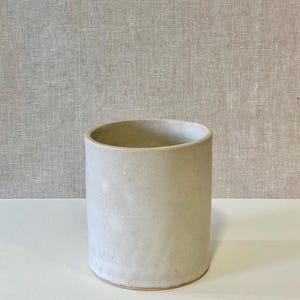 May include: A cylindrical ceramic vase in a neutral, off-white color. The vase has a smooth, matte finish and a simple, minimalist design. The interior is a slightly darker shade of the same color. The vase is suitable for home decor.