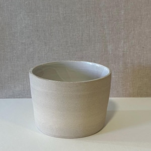 Puede incluir: Una maceta cilíndrica de cerámica beige con exterior liso e interior blanco. La maceta tiene una base redondeada y una abertura ancha. El color neutro y el diseño sencillo la hacen adecuada para varios estilos de plantas y decoración.