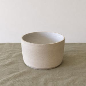 May include: A cylindrical ceramic bowl with a matte, light beige exterior and a smooth, glossy white interior. The bowl features straight, vertical sides and a flat base, showcasing a clean, simple design with visible clay texture.