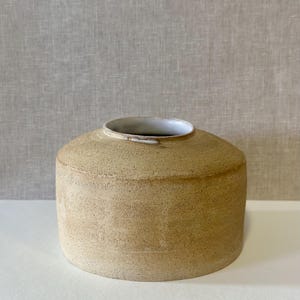 Puede incluir: Un jarrón cilíndrico de cerámica hecho a mano, con un cuerpo ancho y redondeado y una abertura estrecha. El jarrón tiene un exterior texturizado de color beige y un borde interior blanco. La paleta de colores neutros y el diseño minimalista lo hacen adecuado para varios estilos de decoración.