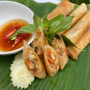 May include: A plate of crispy spring rolls with a side of dipping sauce and a cucumber slice. The rolls are filled with vegetables and meat and are arranged on a green banana leaf.