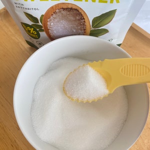 A white bowl filled with white granulated sugar. A yellow plastic spoon with a white handle is scooping up the sugar. The spoon is filled with sugar. The bowl is sitting on a wooden surface. The background shows a bag of monk fruit sweetener with the words 'Monk Fruit Sweetener' and '0% Calories' printed on it.