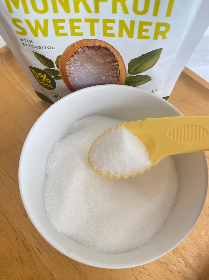 A white bowl filled with white granulated sugar. A yellow plastic spoon with a white handle is scooping up the sugar. The spoon is filled with sugar. The bowl is sitting on a wooden surface. The background shows a bag of monk fruit sweetener with the words 'Monk Fruit Sweetener' and '0% Calories' printed on it.