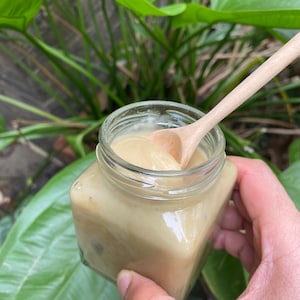 May include: A glass jar filled with a light brown, creamy substance. A wooden spoon is partially submerged in the jar, with the handle extending out of the jar.