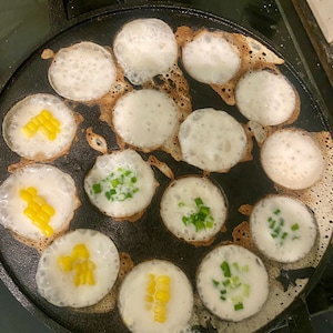 May include: A black cast iron pan with 15 round molds filled with a white batter. Some of the molds have yellow corn kernels and some have green onions on top.