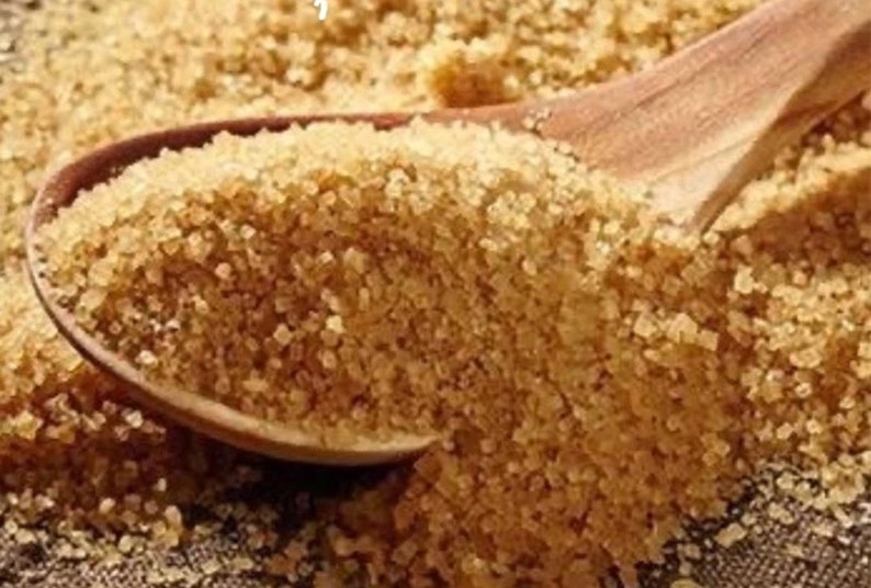 May include: A close-up of brown granulated sugar in a wooden spoon. The sugar is a light brown color and has a grainy texture.