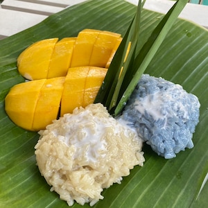 Puede incluir: Un plato de arroz pegajoso con mango. El arroz es blanco y azul, y el mango es amarillo. La comida se sirve en una hoja de plátano verde.