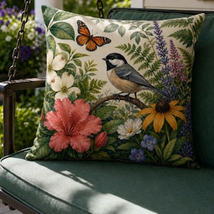 May include: Square throw pillow with a botanical design featuring a bird perched on a branch, surrounded by flowers and a butterfly. The pillow has a green border and is set on a green swing.