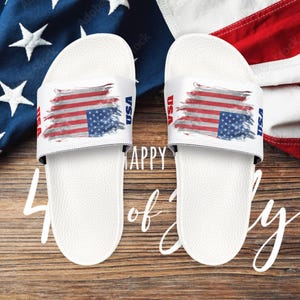 May include: Pair of white slide sandals featuring a painted American flag design on the strap. The word "USA" is printed on the side of the strap. The sandals are displayed on a wooden surface with a patriotic theme.