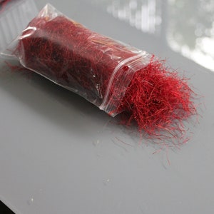 May include: A clear plastic bag filled with vibrant red saffron threads. The bag is partially open, with some threads spilling out onto a gray surface. The saffron threads are long and thin, with a deep red color.