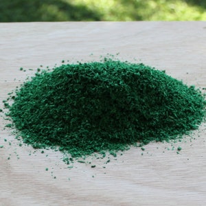 May include: A pile of finely shredded, vibrant green material sits on a light-colored wooden surface. The texture of the material appears granular and uniform, creating a dense, textured mound. The background is blurred, suggesting an outdoor setting.