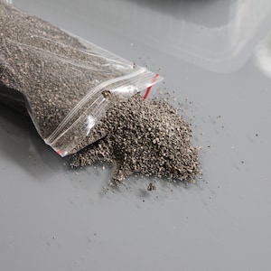 May include: A clear plastic bag partially open, spilling a pile of small, dark gray granules onto a gray surface. The granules are coarse and textured, suggesting a material like gravel or a similar substance.