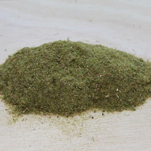 May include: A pile of dried, green herbs, possibly for culinary or medicinal use, sits on a light-colored wooden surface. The herbs appear finely chopped and have a natural, earthy texture.