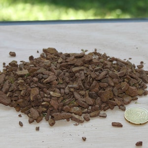 May include: A pile of brown, irregularly shaped wood chips, possibly mulch, sits on a light-colored wooden surface. A 20-cent coin is next to the wood chips for size comparison. The background is blurred.