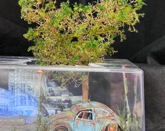 Op de afbeelding: Een miniatuur diorama met een verweerde, blauwe oldtimer, gedeeltelijk verpletterd, in een doorzichtige vitrine. De scène bevat een boom, gebladerte en een stadsgezicht op de achtergrond, wat een post-apocalyptisch thema creëert.