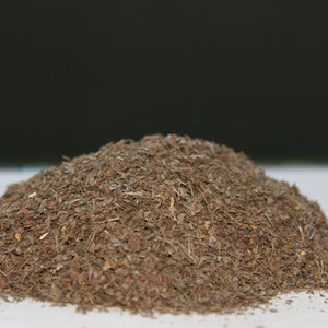 May include: A pile of dried, brown, and tan herbs or spices. The texture appears coarse and granular, with visible small pieces and fragments. The background is a dark, solid color, contrasting with the lighter tones of the herbs.