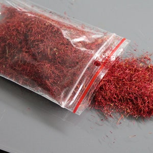 May include: A clear plastic bag filled with vibrant red saffron threads, with some threads spilling out onto a gray surface. The saffron is a deep, rich red color, and the bag has a red zipper closure.