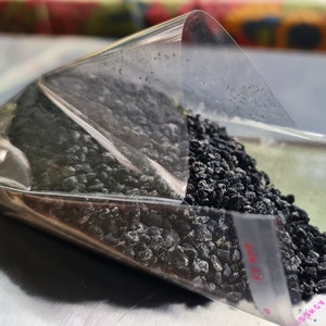 May include: A clear plastic bag filled with small, dark, granular material. The bag is partially open, revealing the contents. The bag is resting on a silver surface, possibly a scale. The words "REMOVE LINER" are printed on the bag.