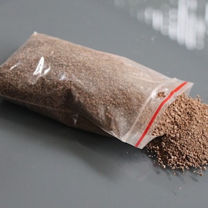 May include: A clear plastic bag, partially open, filled with small, brown granular material. Some of the material has spilled out onto a gray surface. The bag has a red stripe along the opening.