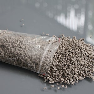 May include: A clear plastic bag partially open, filled with small, gray, granular material. The granules are scattered on a gray surface. The bag is sealed with a red and white zipper. The image is well-lit, showing the texture and color of the material.