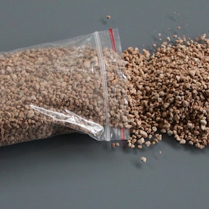 May include: A clear plastic bag partially filled with small, light brown, pebble-like material, with a red zipper closure. Some of the material is spilled out onto a gray surface, forming a small pile.