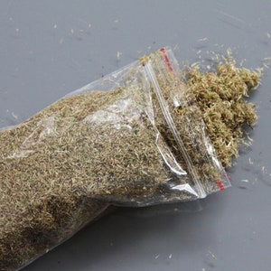 May include: A clear plastic bag filled with dried herbs, partially open, spilling onto a gray surface. The herbs are a mix of green and brown, with a fine, shredded texture. The bag has a red and white zipper.
