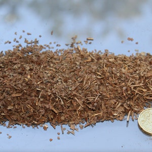 May include: A pile of shredded, dried tobacco leaves, in shades of brown, with a 20 Euro cent coin for scale. The tobacco appears to be finely cut, with visible stems and leaf fragments. The coin is gold-colored.