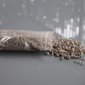 May include: A clear plastic bag filled with small, gray, pebble-like stones, partially open, with a pile of the same stones spilled out onto a gray surface. The stones have a rough texture and varying shades of gray.