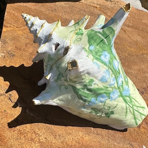 May include: A decorative seashell painted with a light green and white base, adorned with blue floral patterns and gold accents. The shell's exterior features a textured surface and a pointed spiral tip.