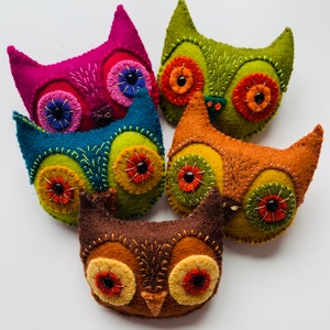 Puede incluir: Cinco adornos de búho de fieltro en varios colores, incluyendo rosa, azul verdoso, verde, marrón y naranja. Cada búho tiene detalles bordados y ojos de botón negros.