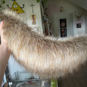 Op de afbeelding: Een lang, gebogen staartaccessoire van imitatiebont in tinten bruin, beige en zandkleur. De pluche, ruige textuur imiteert dierenvacht en is voorzien van een dunne, verstelbare bruine lederachtige riem met een metalen gesp voor bevestiging.