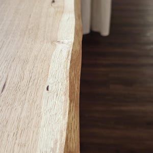 May include: A long, rectangular wooden shelf or tabletop featuring a natural live edge with an irregular, organic contour. The surface displays a light tan wood grain with visible knots and a smooth, unfinished texture along the length.