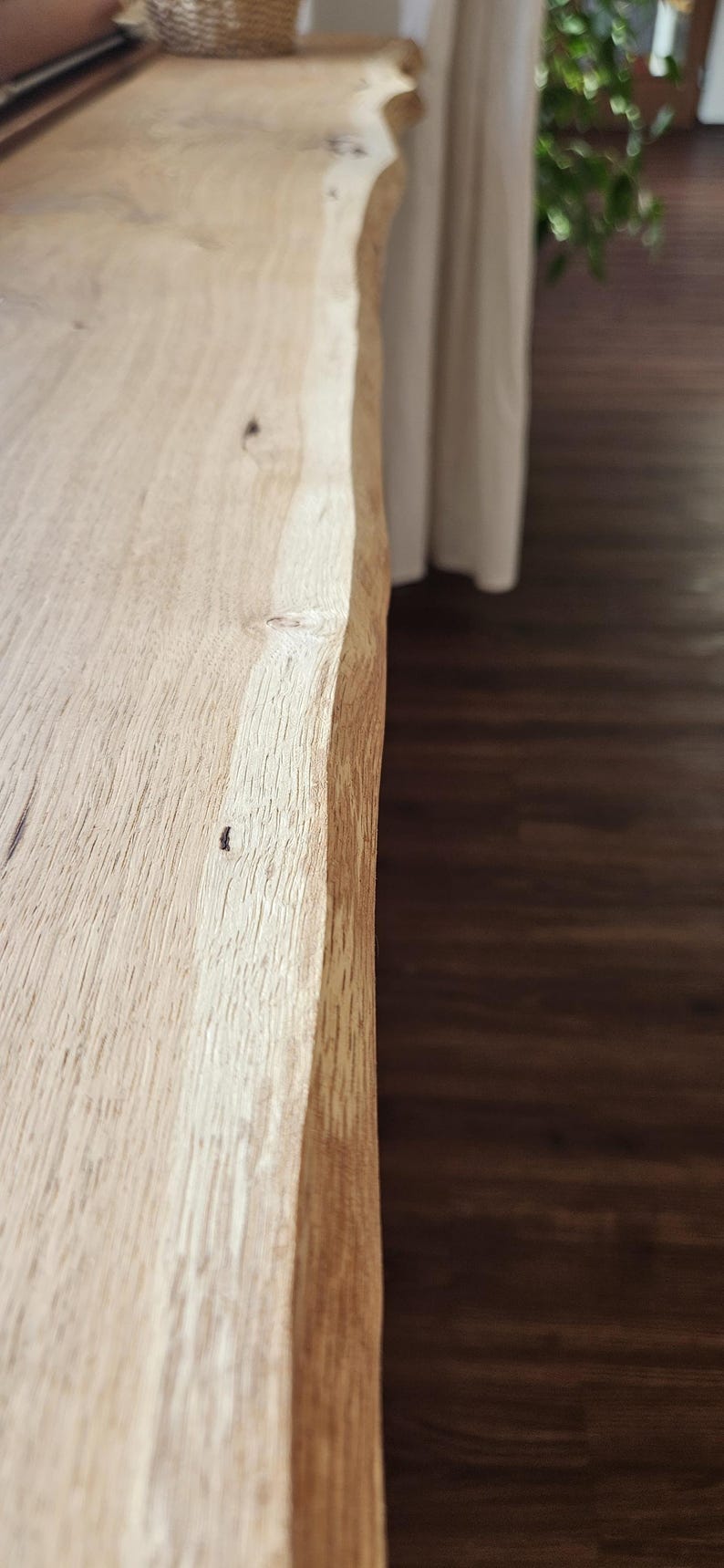 May include: A long, rectangular wooden shelf or tabletop featuring a natural live edge with an irregular, organic contour. The surface displays a light tan wood grain with visible knots and a smooth, unfinished texture along the length.