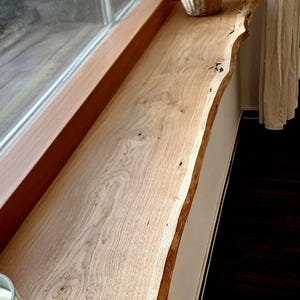 May include: A long, rectangular wooden window sill featuring a natural live edge with visible wood grain, knots, and light brown tones. The piece is installed horizontally along a window frame, with a woven vase and a glass candle holder present.