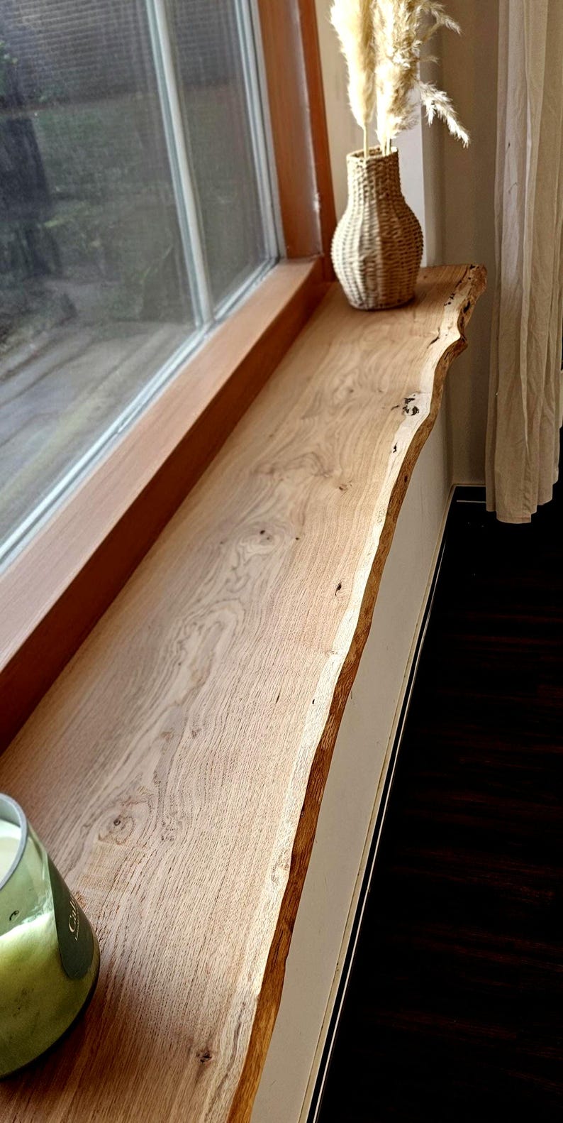 May include: A long, rectangular wooden window sill featuring a natural live edge with visible wood grain, knots, and light brown tones. The piece is installed horizontally along a window frame, with a woven vase and a glass candle holder present.