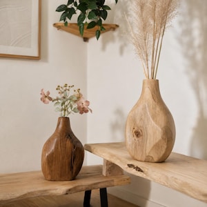 Pode incluir: Dois vasos de madeira de formas orgânicas com superfícies lisas e polidas. O vaso mais pequeno, de cor castanho-escuro, contém um ramo de pequenas flores cor-de-rosa, enquanto o vaso maior, de cor bege claro, contém hastes de erva-das-pampas.