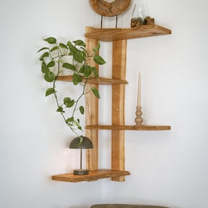 Op de afbeelding: Houten hoekplank met vier planken, een decoratieve houten ring en een kleine lamp. Een potplant met hangende groene bladeren en een kaars zijn ook te zien. De planken zijn gemaakt van lichtgekleurd hout.