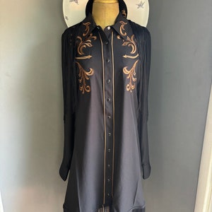 Op de afbeelding: Een zwart lang mouw shirt met gouden borduurwerk en franjes. Het shirt heeft een knoopsluiting en een puntige kraag.