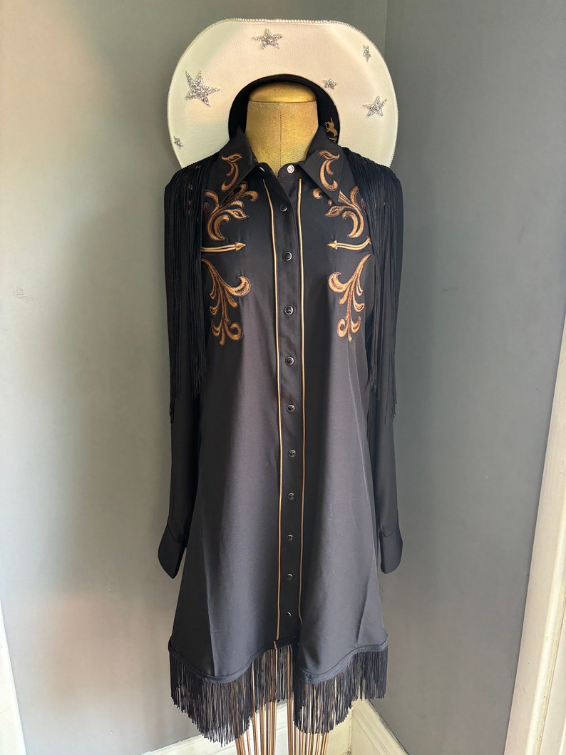 Op de afbeelding: Een zwart lang mouw shirt met gouden borduurwerk en franjes. Het shirt heeft een knoopsluiting en een puntige kraag.