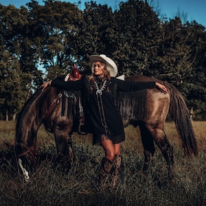 Op de afbeelding: Een vrouw in een zwart jasje met franjes en een witte cowboyhoed staat naast een bruin paard in een veld. Ze leunt tegen de rug van het paard en heeft haar armen uitgestrekt.