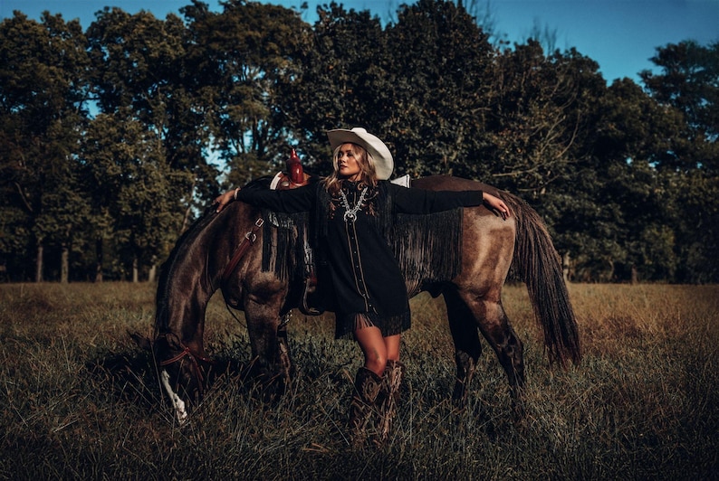 Op de afbeelding: Een vrouw in een zwart jasje met franjes en een witte cowboyhoed staat naast een bruin paard in een veld. Ze leunt tegen de rug van het paard en heeft haar armen uitgestrekt.