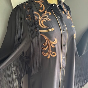 Op de afbeelding: Zwart langarm shirt met gouden borduurwerk en franjes. Het shirt heeft een knoopsluiting en een puntige kraag.