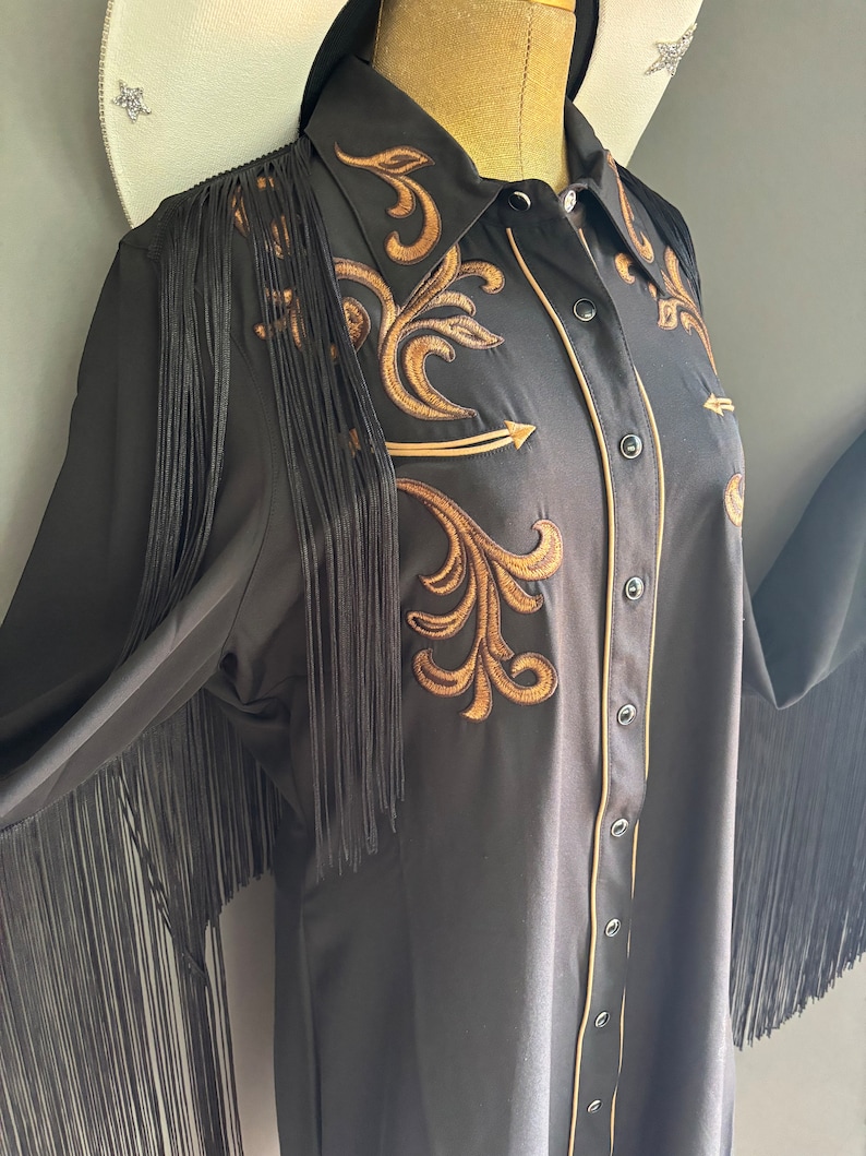 Op de afbeelding: Zwart langarm shirt met gouden borduurwerk en franjes. Het shirt heeft een knoopsluiting en een puntige kraag.