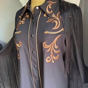 Op de afbeelding: Een zwart lang mouw shirt met gouden borduurwerk en zwarte franjes. Het shirt heeft een knoopsluiting en een puntige kraag.