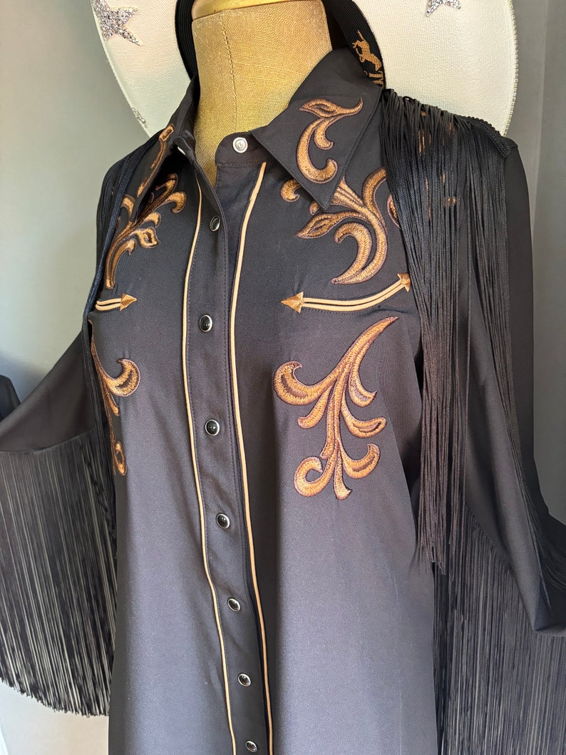 Op de afbeelding: Een zwart lang mouw shirt met gouden borduurwerk en zwarte franjes. Het shirt heeft een knoopsluiting en een puntige kraag.