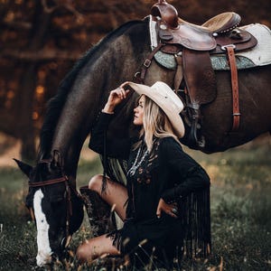 Op de afbeelding: Een vrouw met een witte cowboyhoed en een zwarte jurk met franjes zit naast een bruin paard met een zadel. Het paard graast op groen gras.