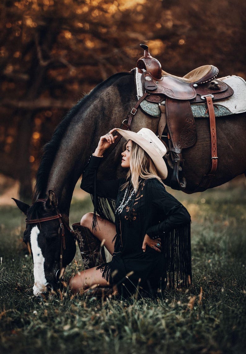 Op de afbeelding: Een vrouw met een witte cowboyhoed en een zwarte jurk met franjes zit naast een bruin paard met een zadel. Het paard graast op groen gras.