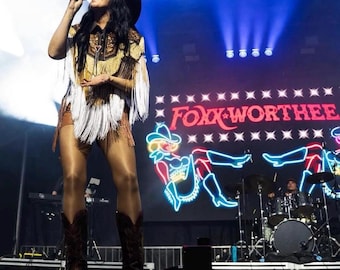 May include: A performer wearing a black cowboy hat, a tan and brown western-style shirt with long white fringe, and dark brown cowboy boots. The shirt features intricate embroidery. The stage background displays the neon text FOXX-WORTHEE.