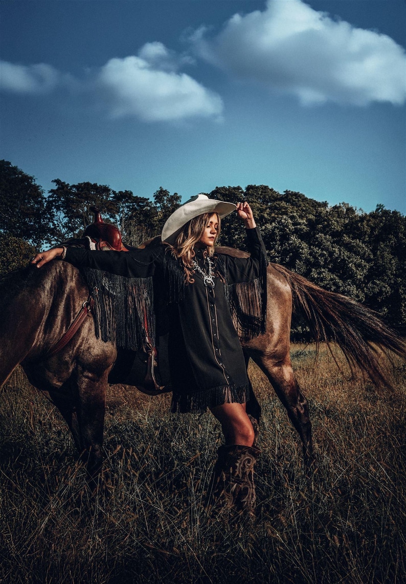 Op de afbeelding: Een vrouw in een zwart shirt met franjes en een witte cowboyhoed staat naast een bruin paard in een veld met hoog gras.